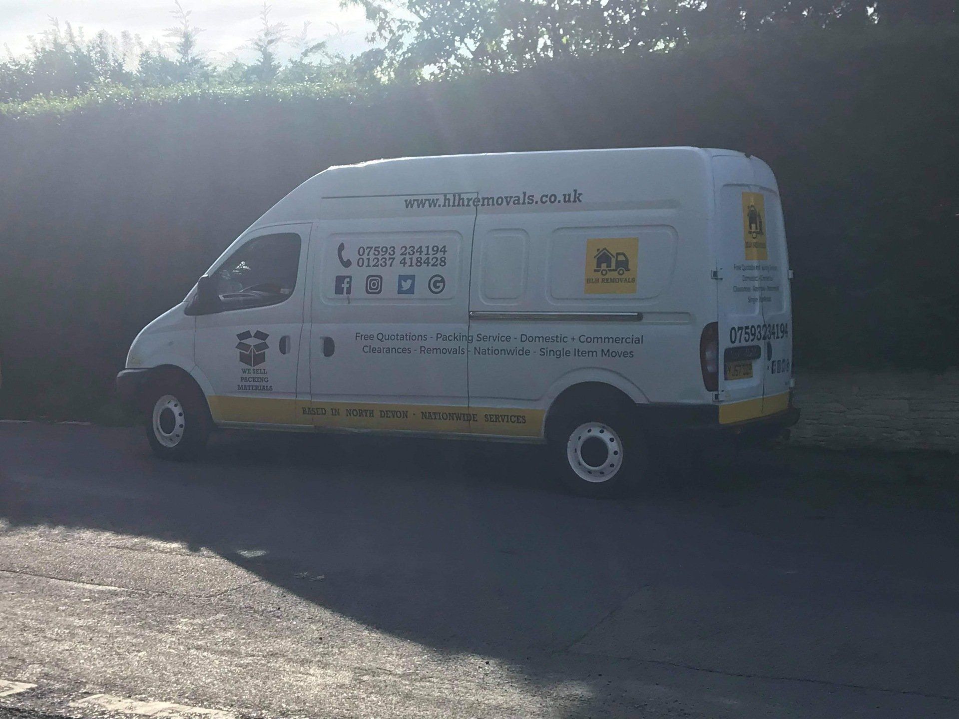 White delivery van parked on a sunny roadside beside a hedge and stone wall.