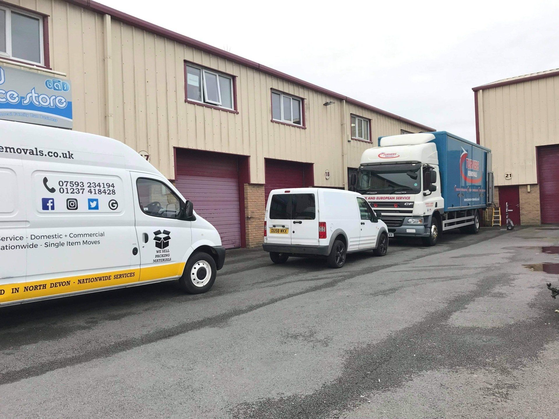Delivery vans and a truck parked outside warehouse loading bays on a paved lot.