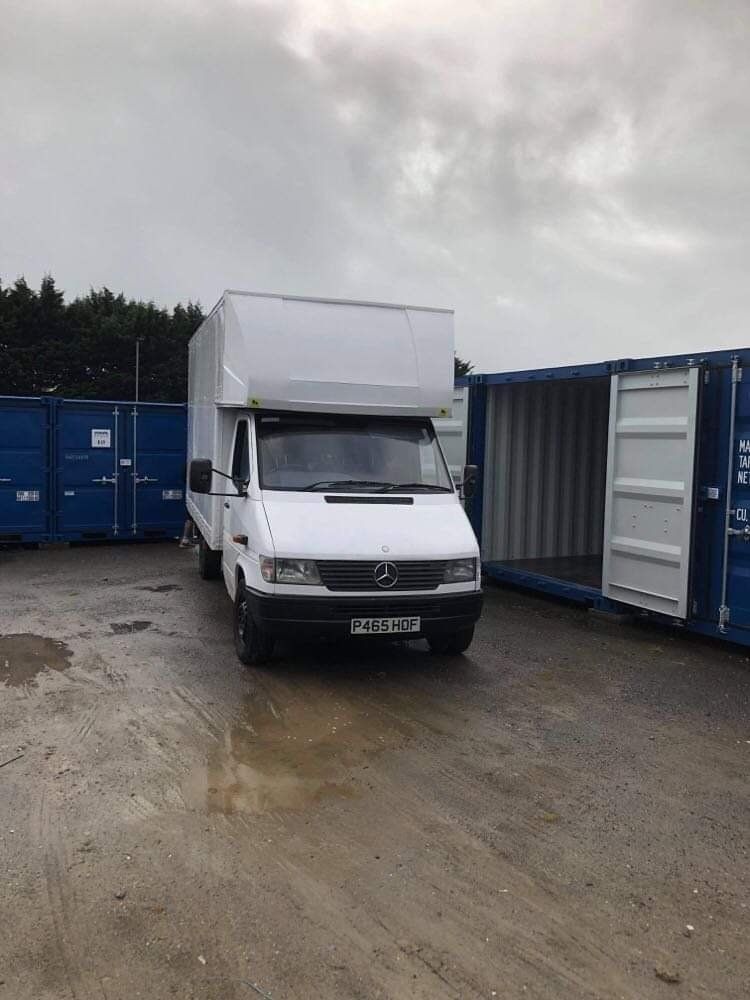 White box truck parked in a lot beside blue shipping containers under a cloudy sky
