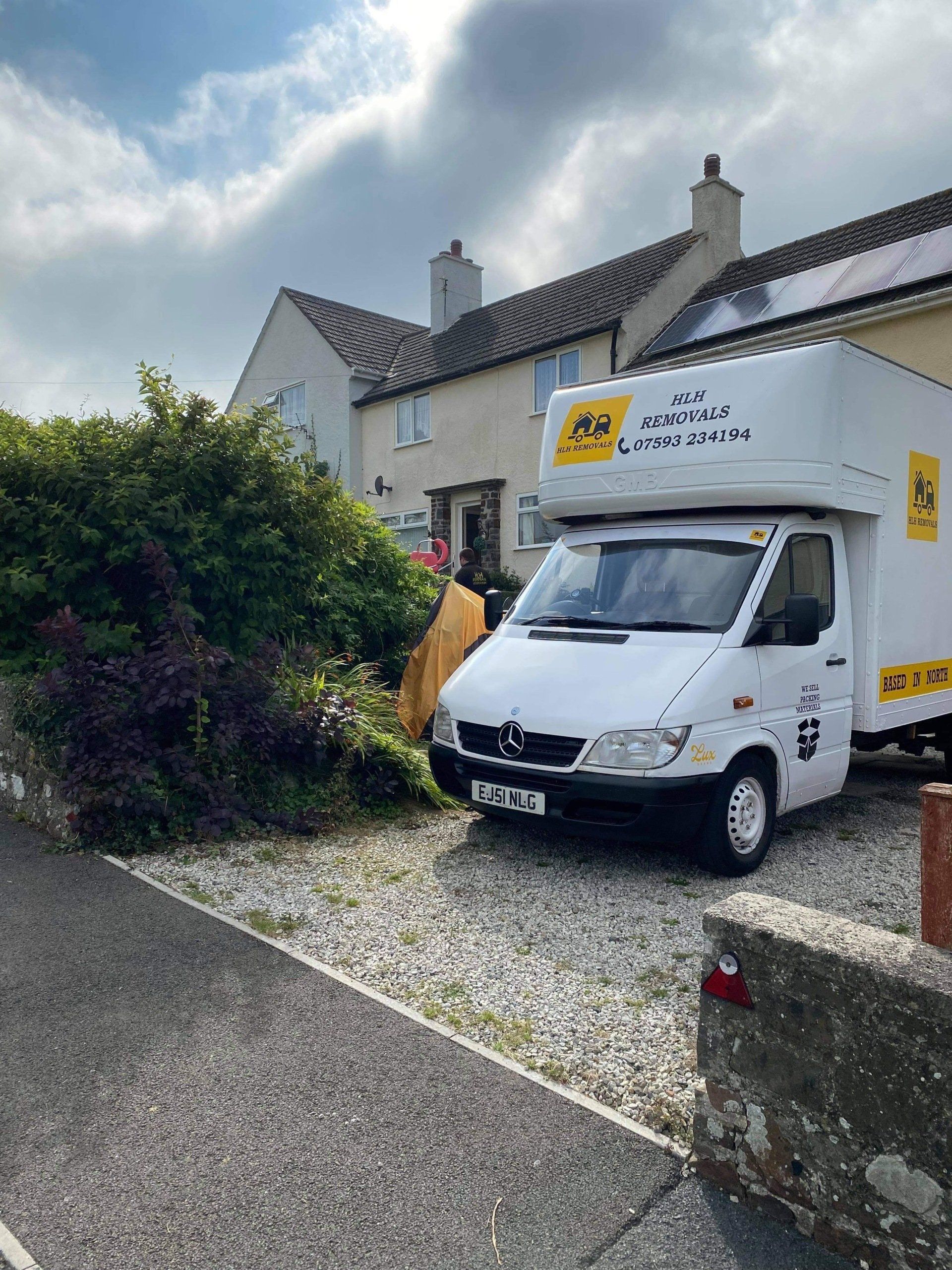White moving van parked in a residential driveway beside hedges and a stone wall, with houses in the background