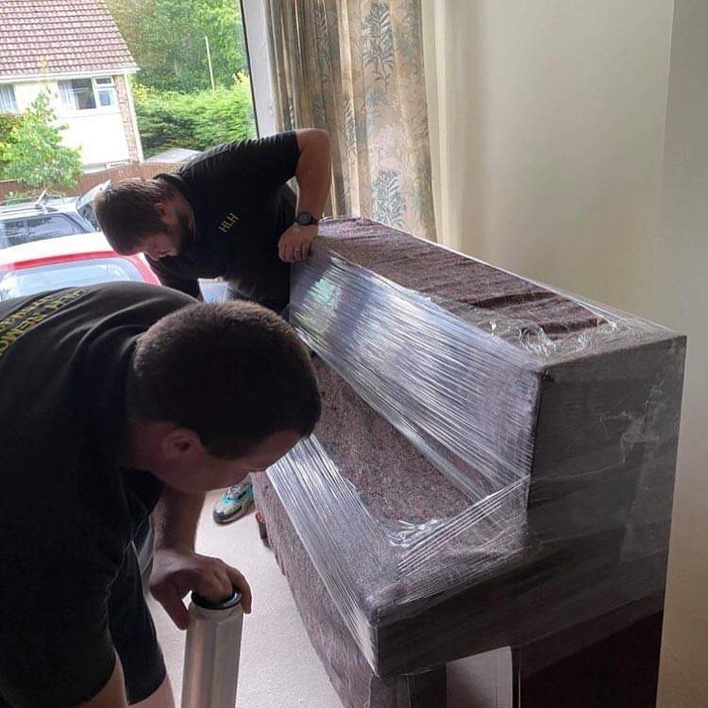 Two people wrapping a large brown wooden item in plastic indoors near a window