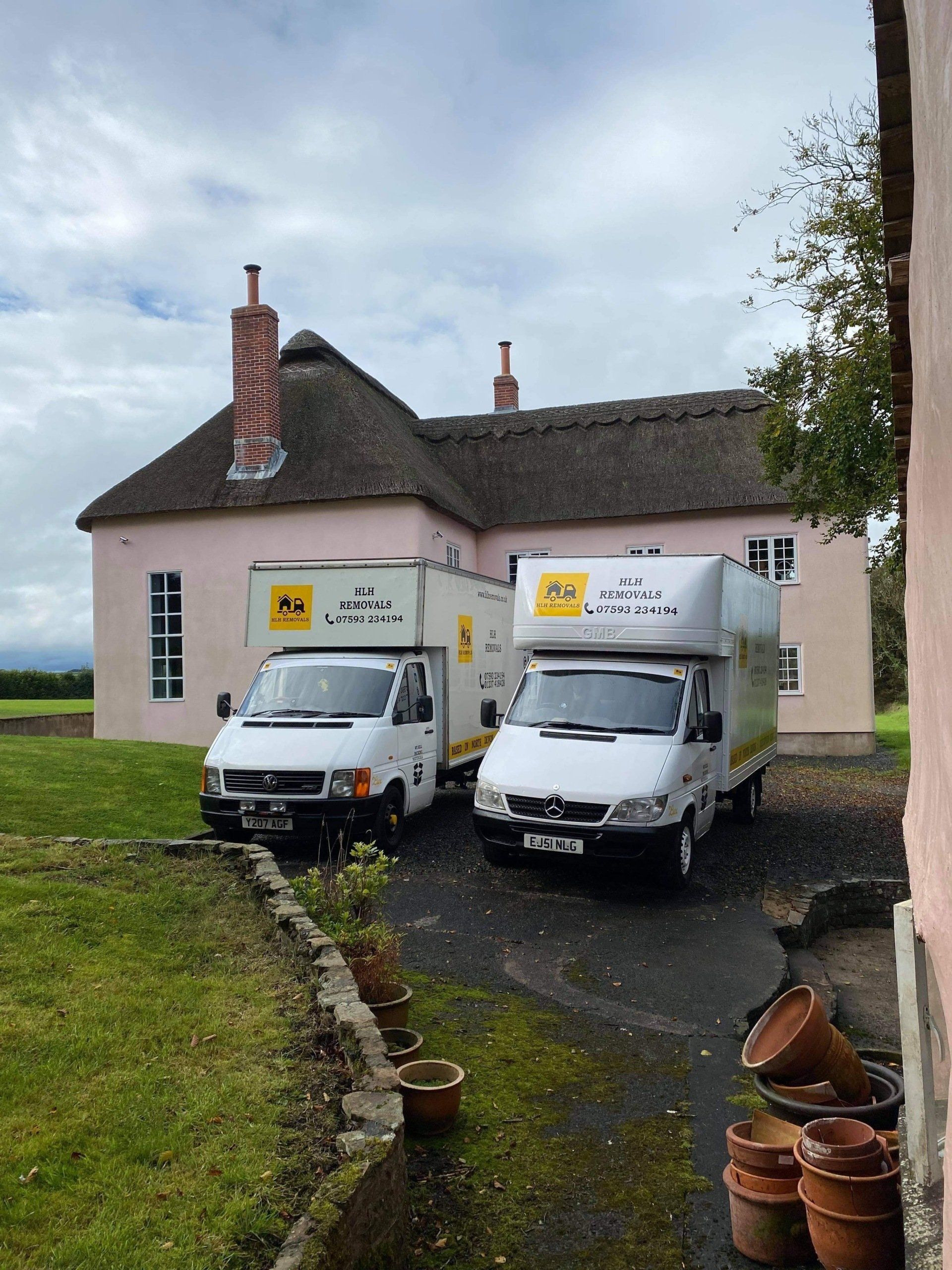 Two delivery vans parked outside a small beige house with a thatched roof on a cloudy day