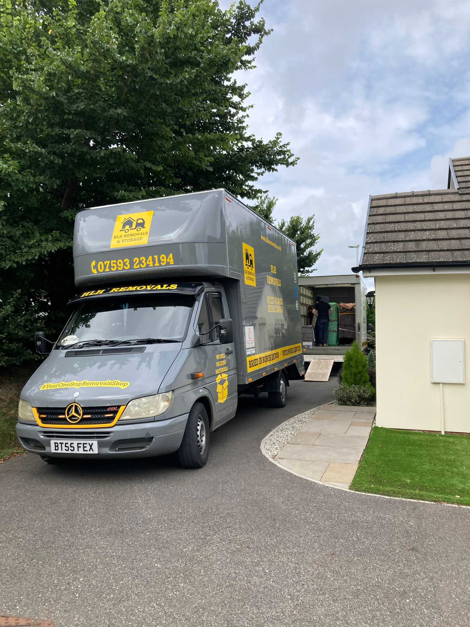 Grey moving truck parked on a driveway beside a house, with yellow company branding and a side ramp.