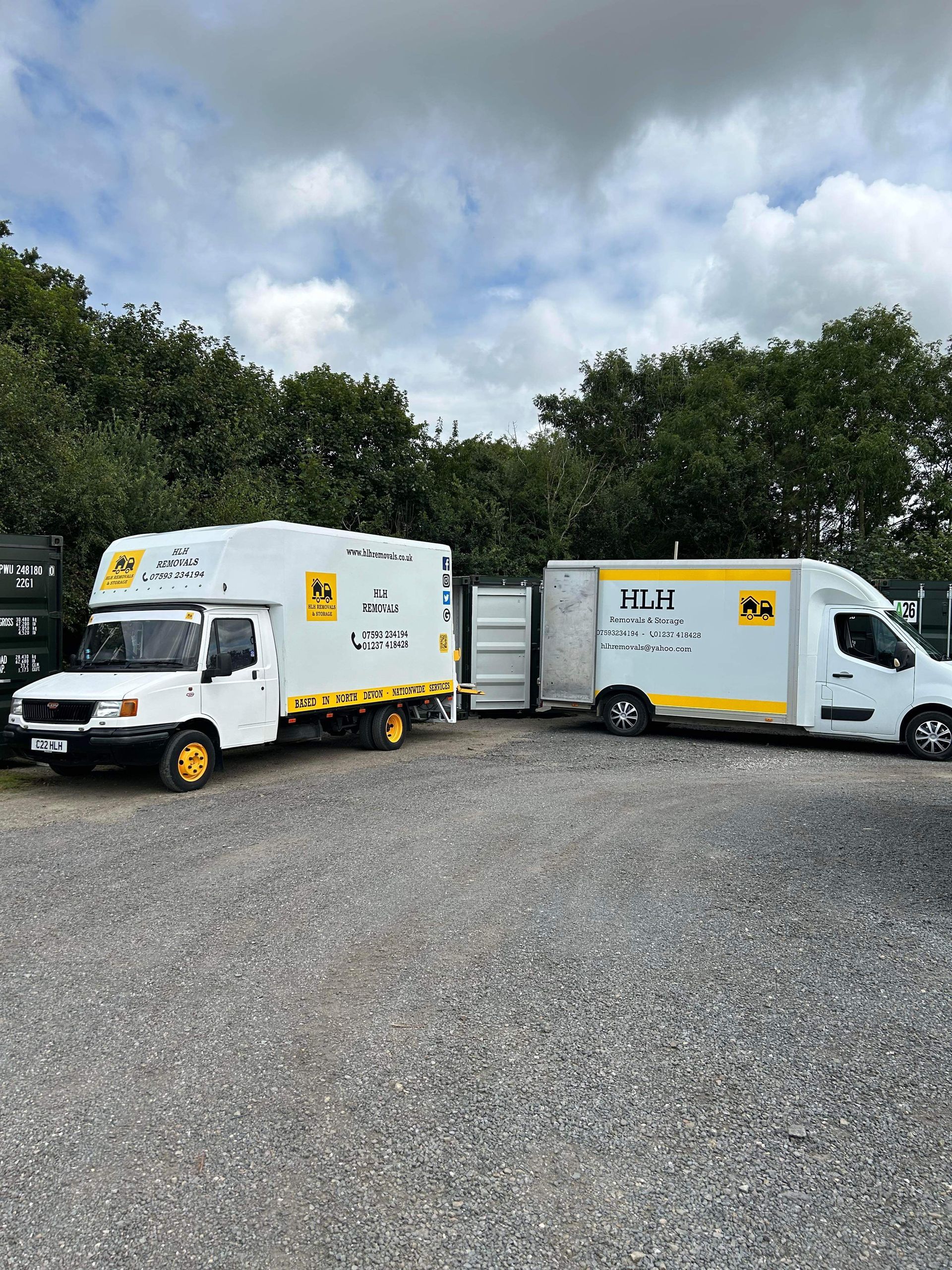 Two white delivery trucks parked on gravel in front of trees and cloudy sky
