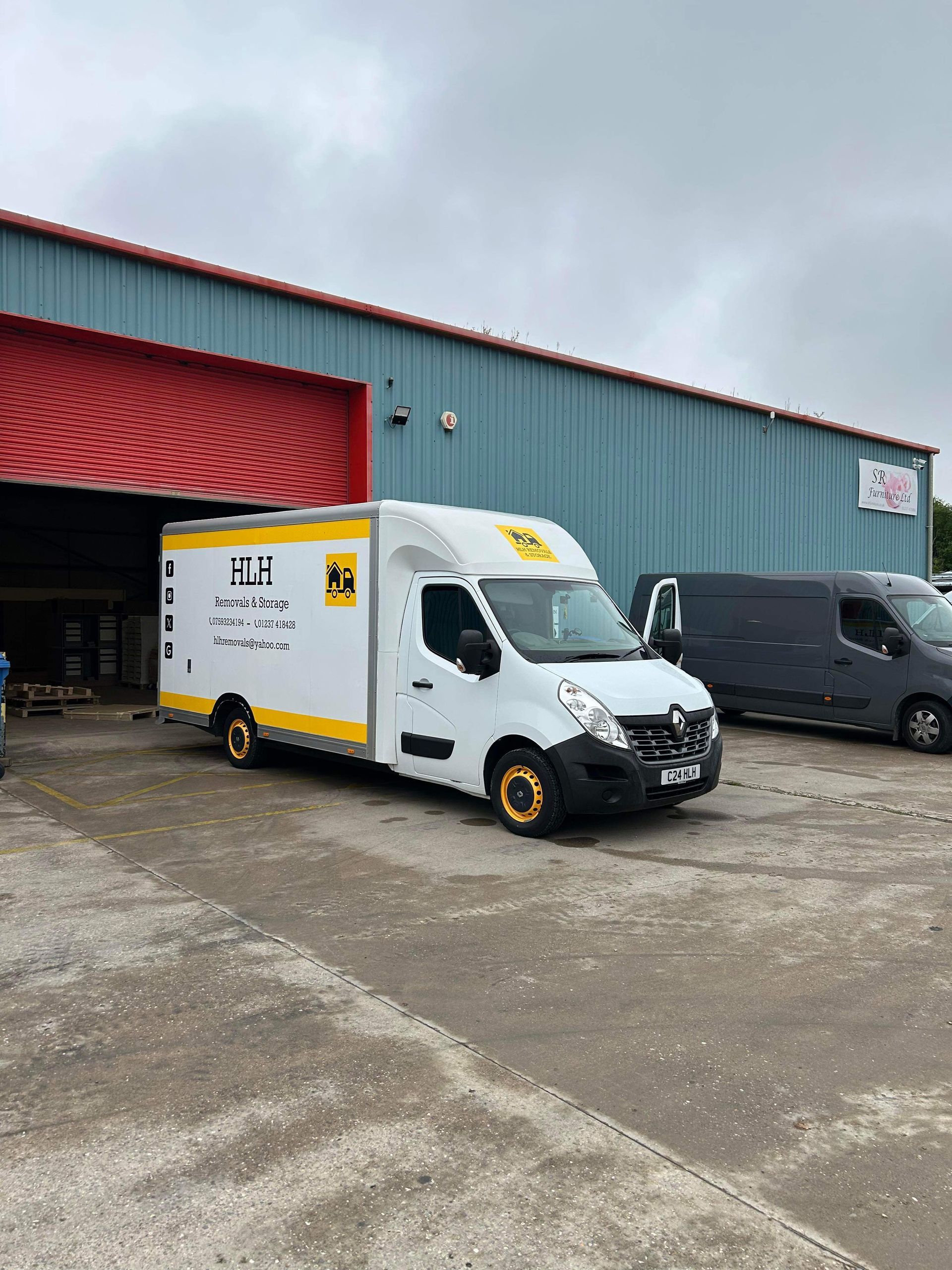 White delivery van parked outside a blue warehouse on a cloudy day, with a gray van nearby.