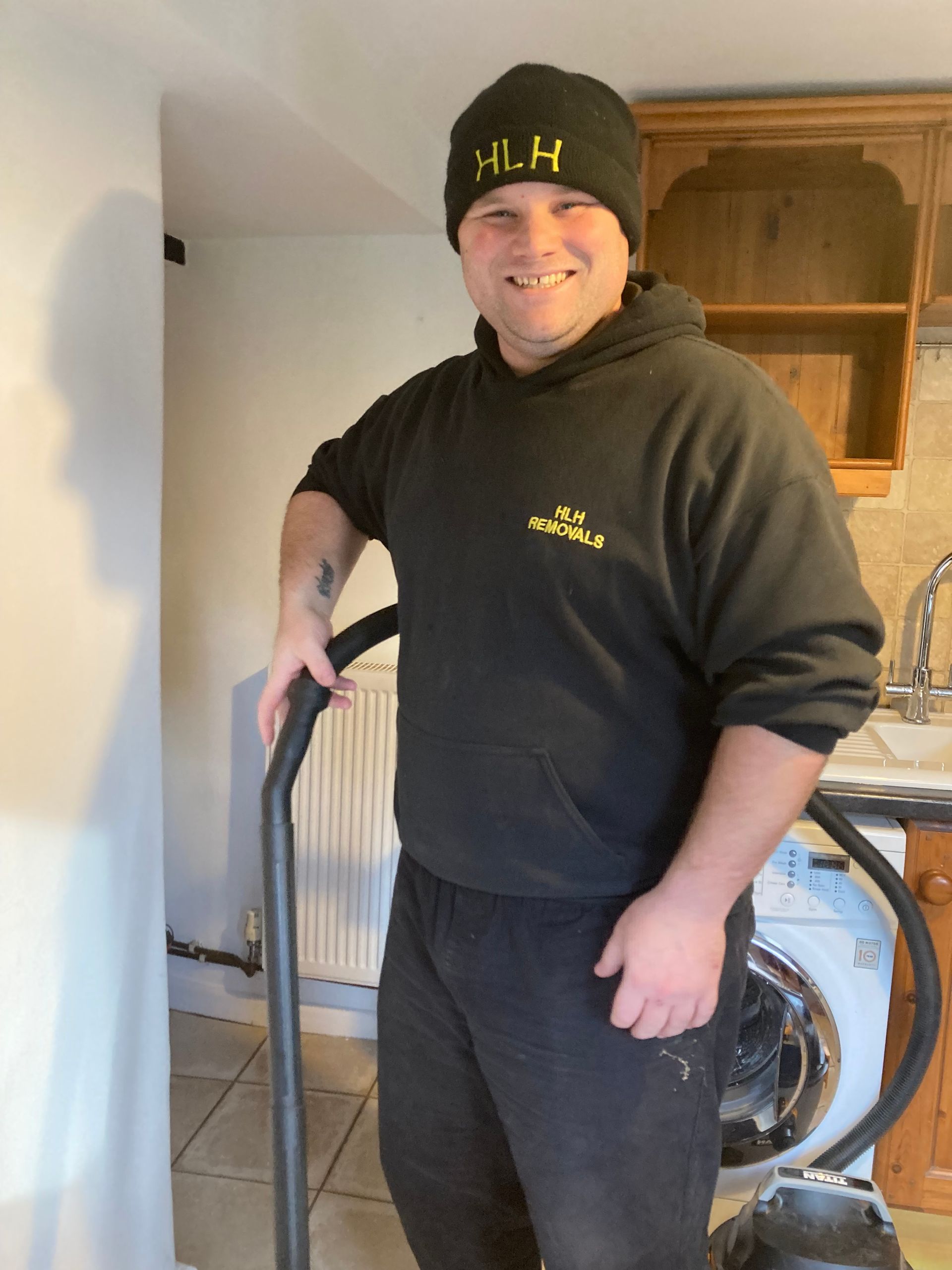 Person in black hoodie and beanie standing indoors beside a vacuum cleaner, smiling.