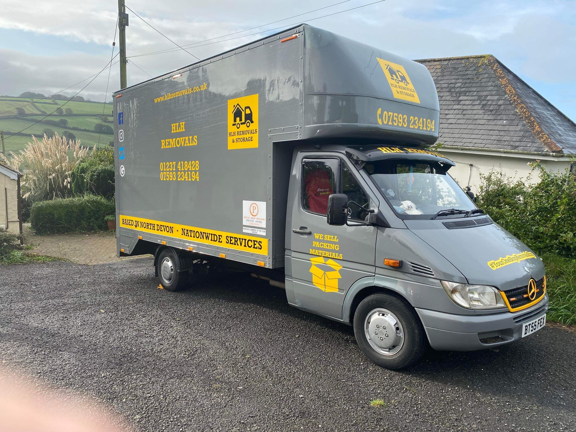 Gray van with yellow signage parked on gravel beside a house in a rural setting