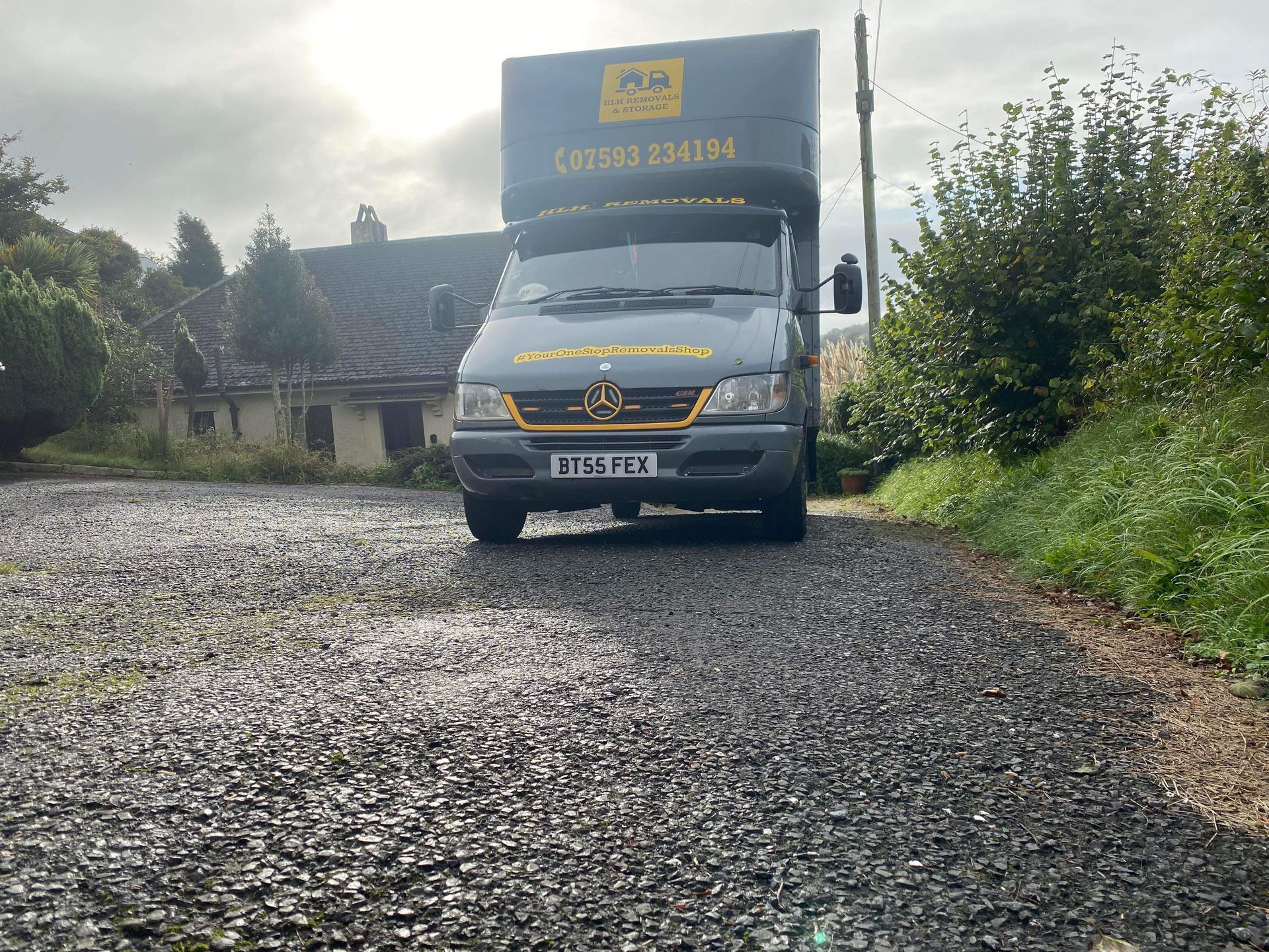 Truck parked on a gravel road beside a house and bushes under a cloudy sky