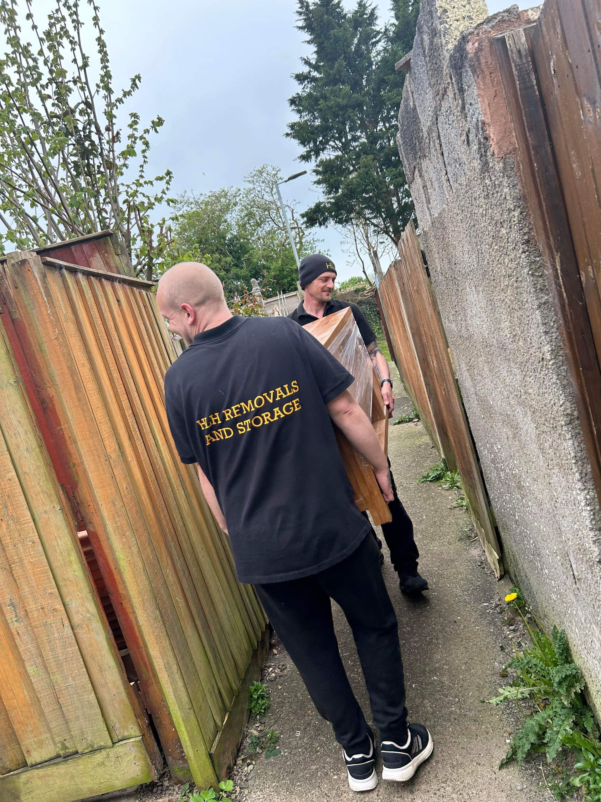 Two people stand in a narrow alley beside a rusty metal fence and stone wall.