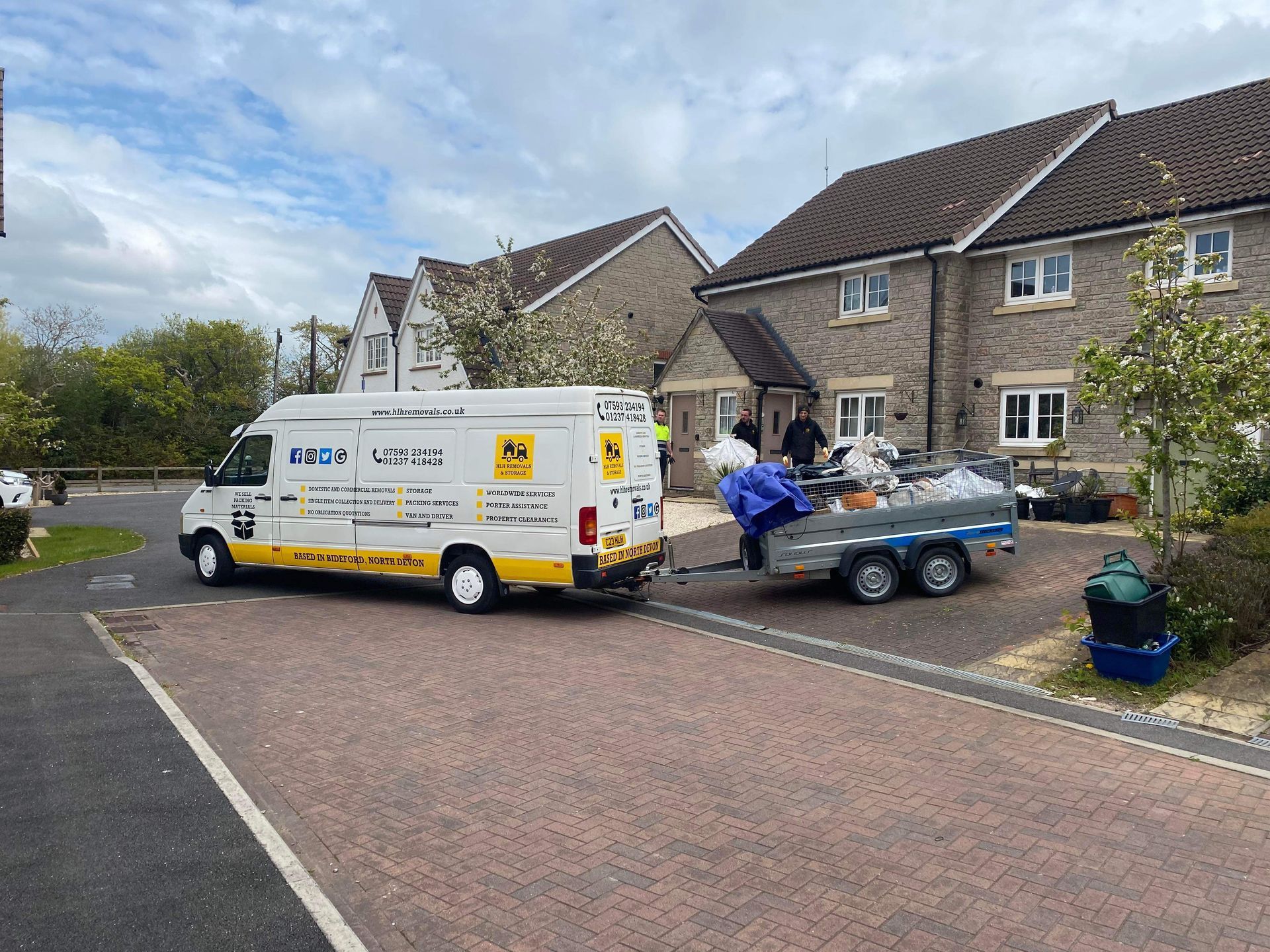 White van and trailer outside stone houses, with workers loading debris in a residential driveway.