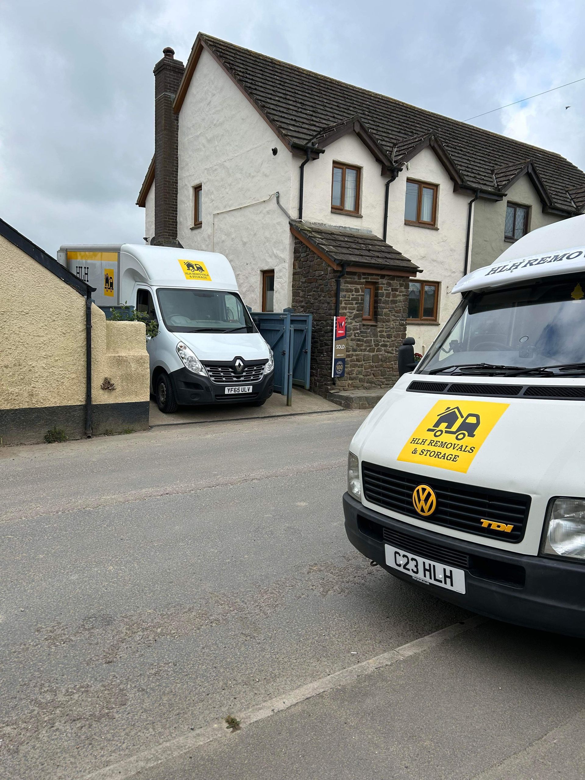 Two white delivery vans parked outside a stone house on a quiet street.