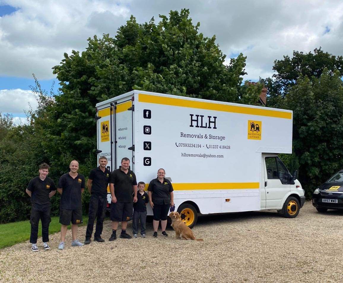 Team standing beside a white-and-yellow HLH box truck in a gravel lot with trees behind them