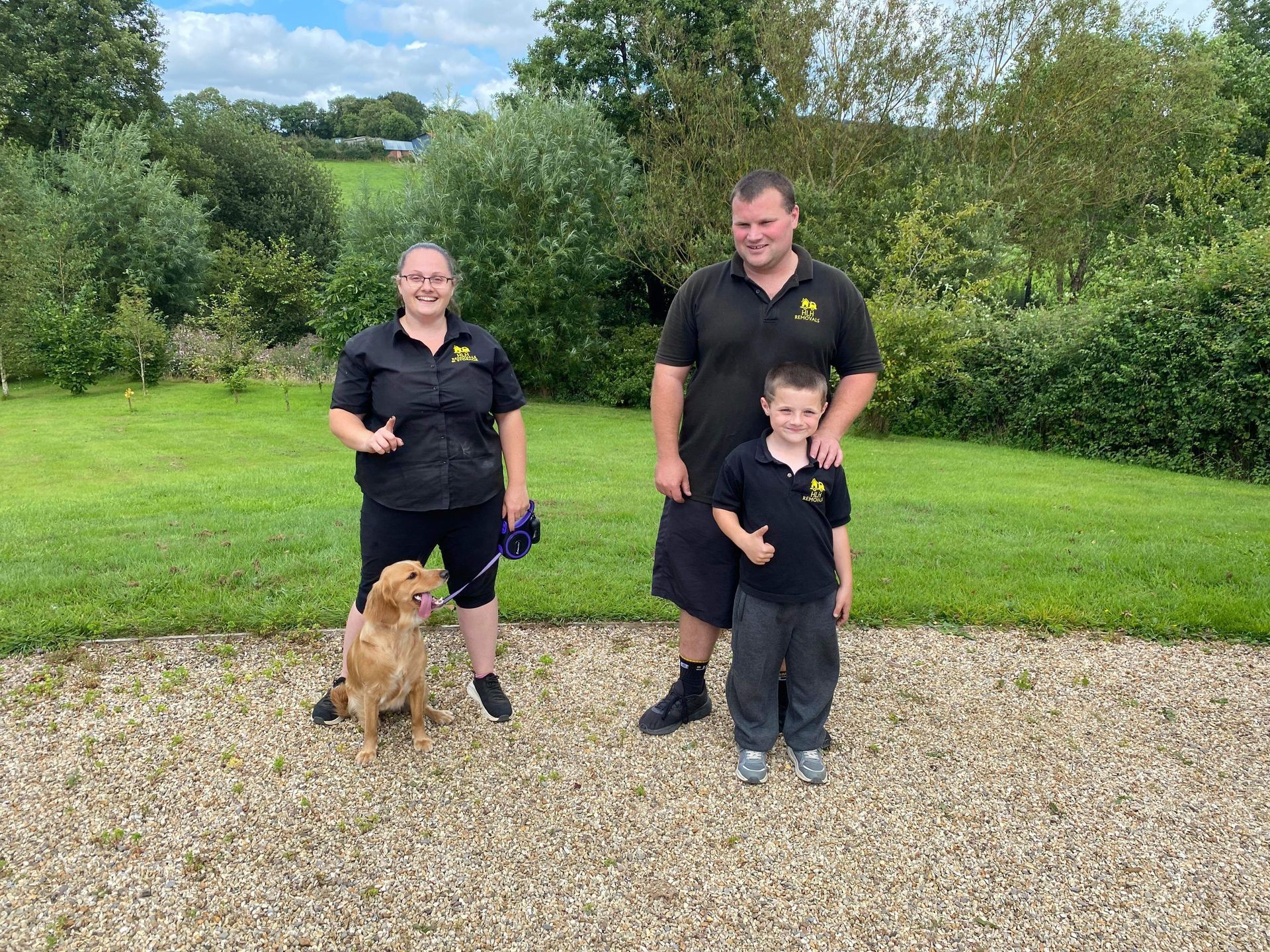 Three people in black shirts standing outdoors with a golden dog on gravel in front of grassy trees.