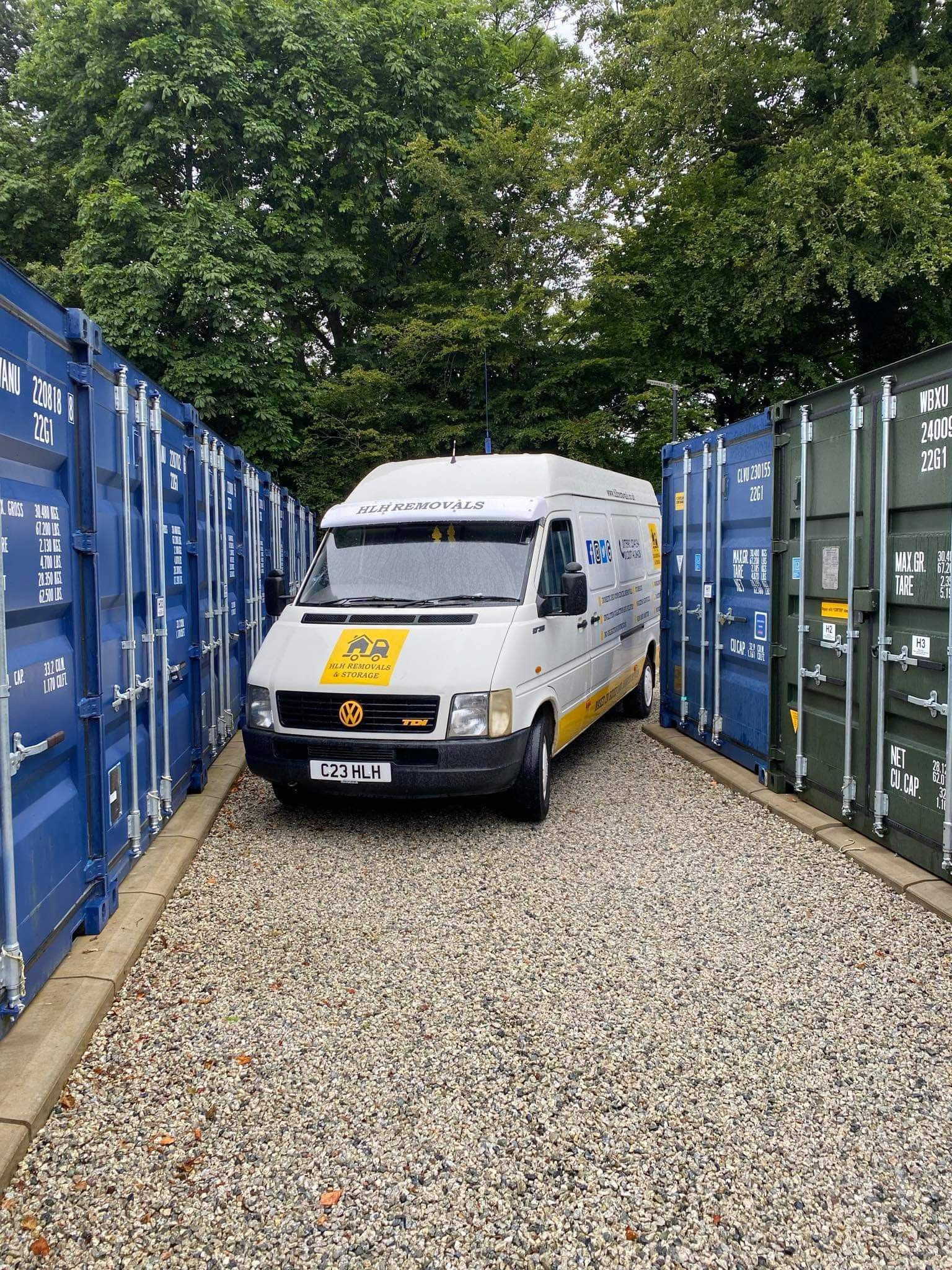 White van parked between blue and green shipping containers on a gravel lane.