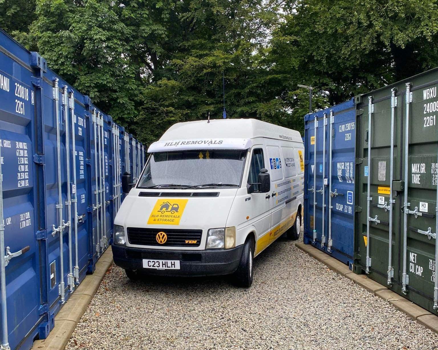 White delivery van parked in a narrow lane between blue and green shipping containers