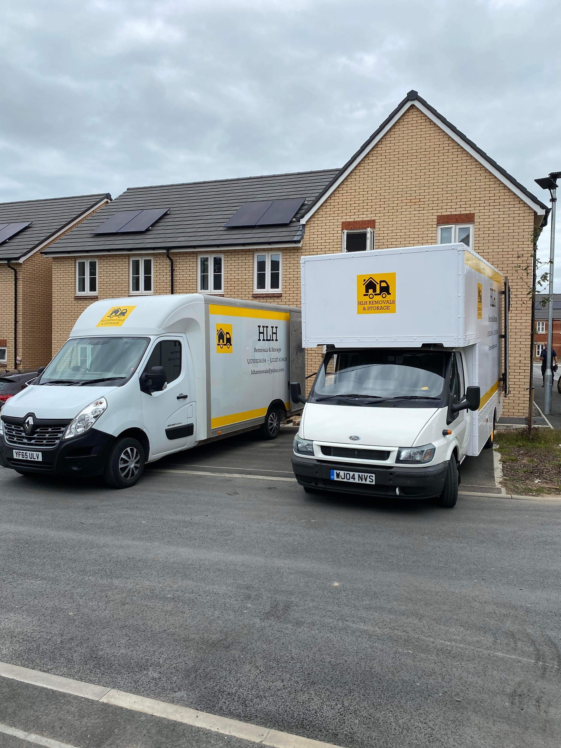 Two white delivery vans parked outside a tan house with a yellow sign on the front.