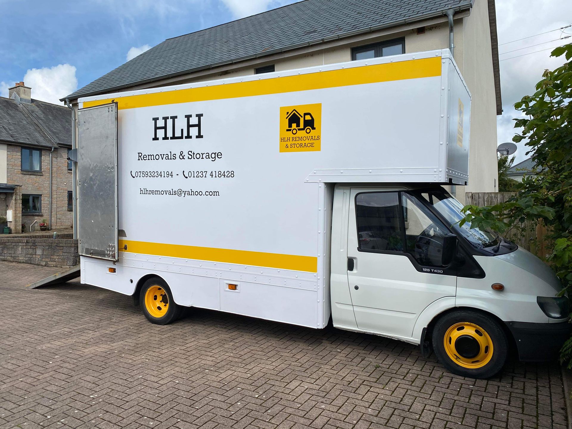 White HLH moving truck with yellow trim parked on a cobblestone street by houses.