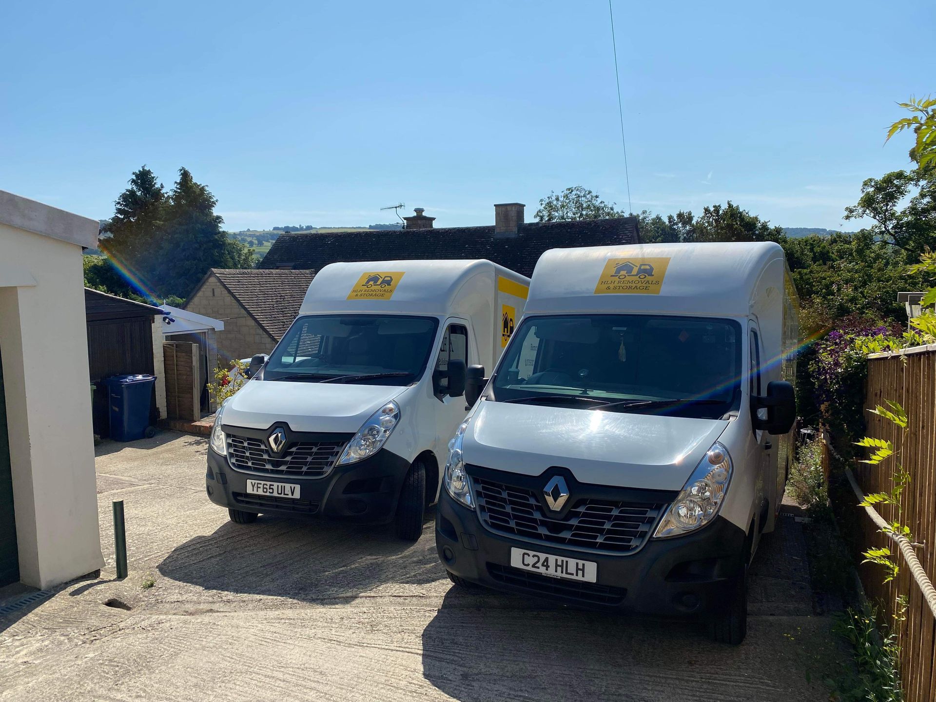 Two white delivery vans parked side by side in a sunny driveway near a house and trees.