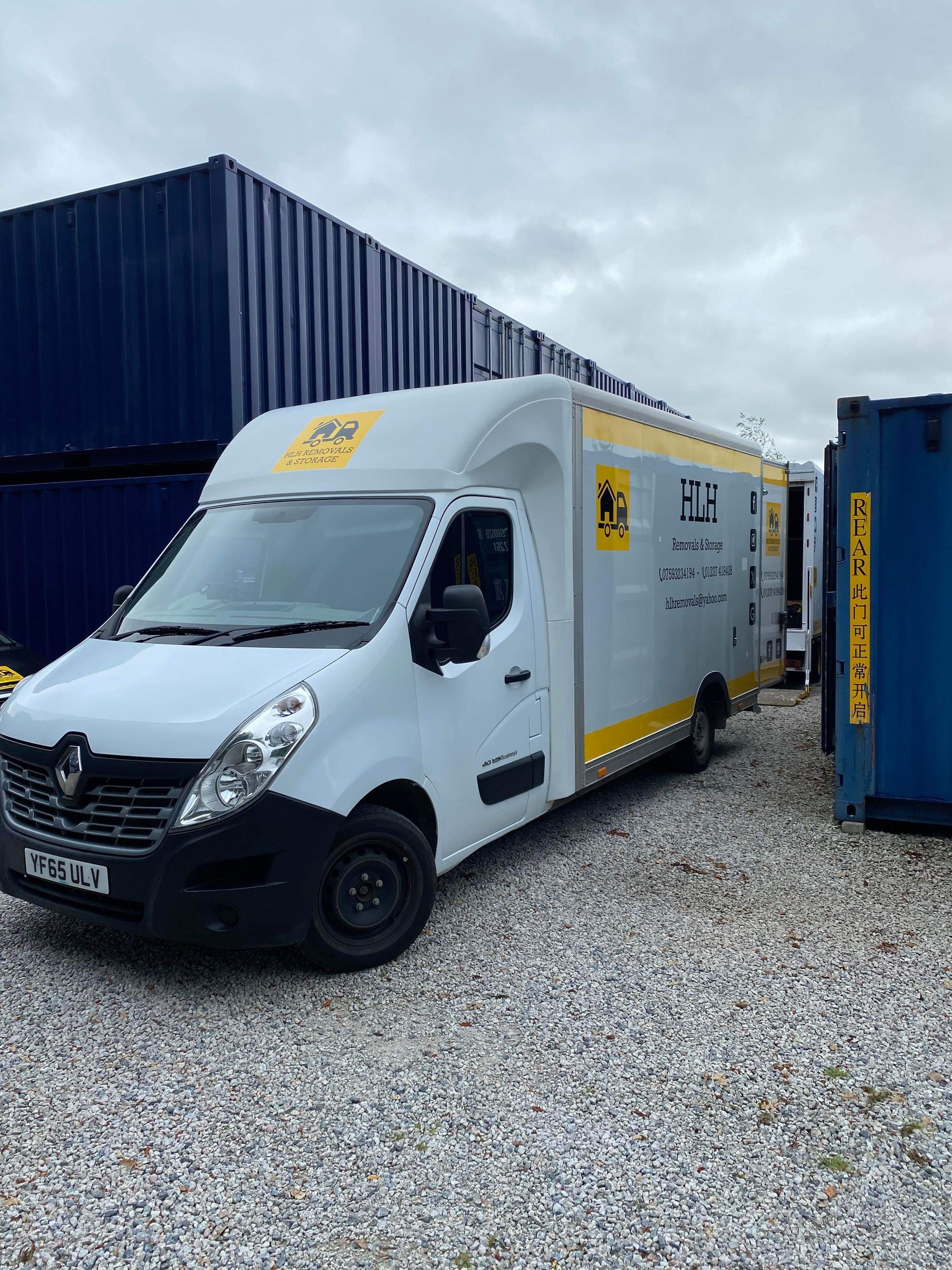 White delivery van with yellow accents parked on gravel beside blue shipping containers