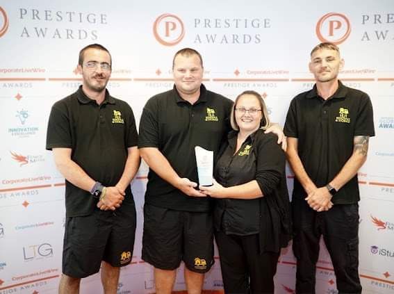 Four people in black uniforms holding an award at a Prestige Awards backdrop