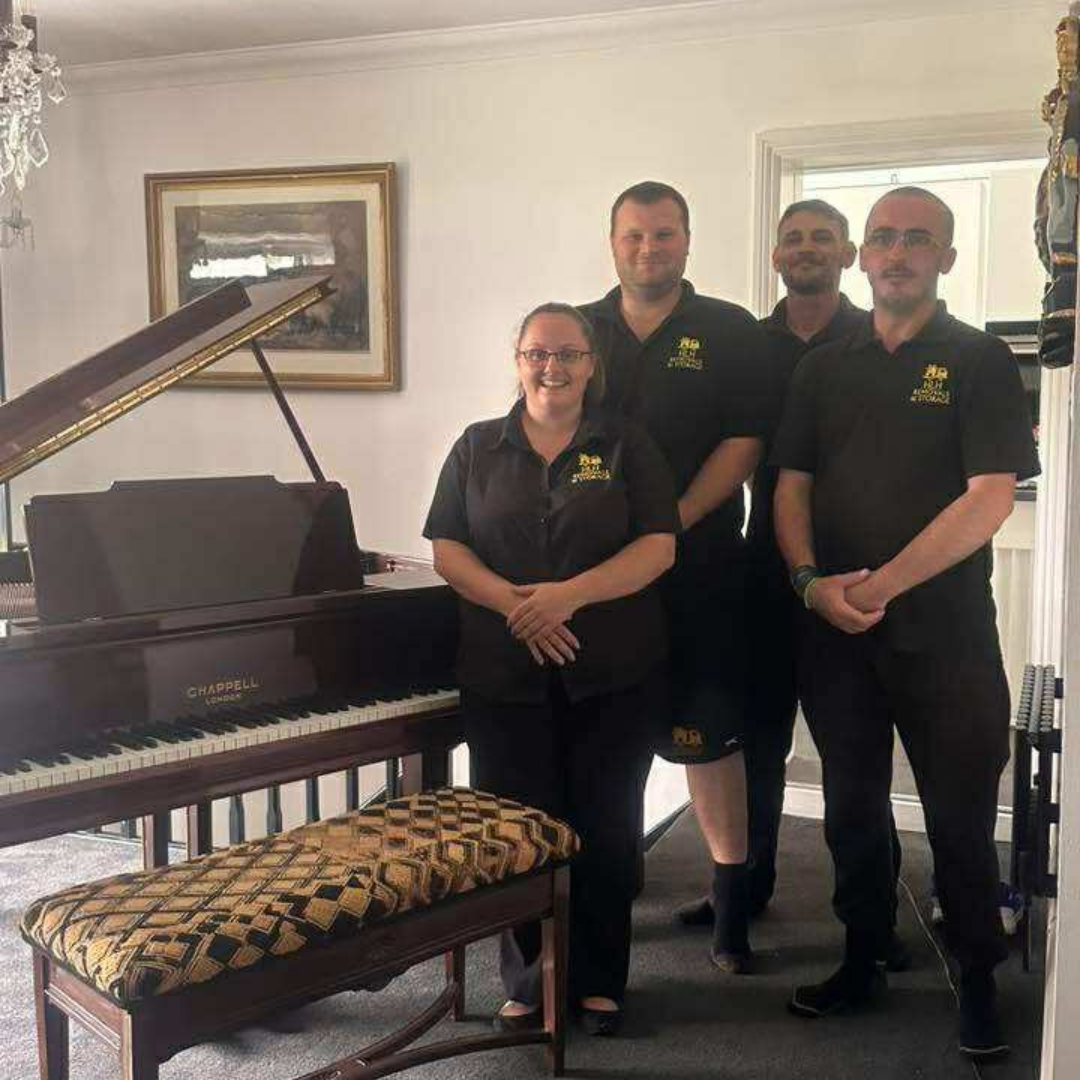 Four people in black shirts stand beside a grand piano in a bright room with a framed painting.