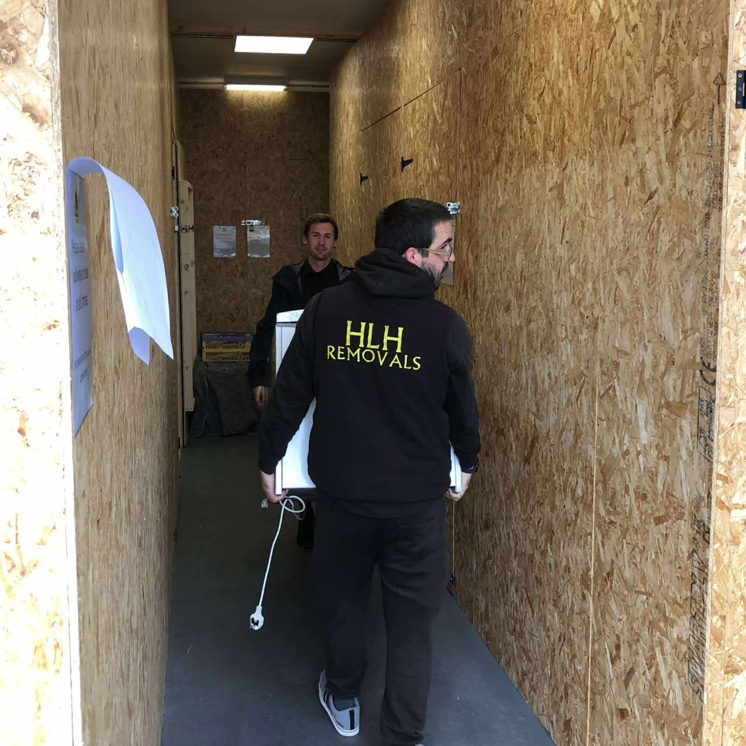 Two movers in black jackets carry items through a narrow plywood-walled hallway.