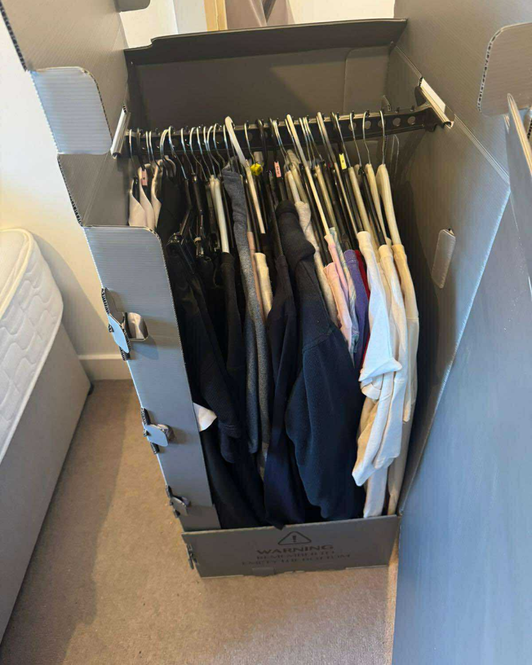 Open cardboard box filled with hanging shirts and jackets in a hallway or room corner