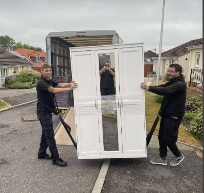 Two workers carrying a white wardrobe on a residential street, with a moving truck behind them.
