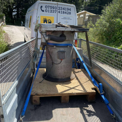 Large metal bin strapped on a flatbed truck, crossing a narrow bridge in a wooded area.