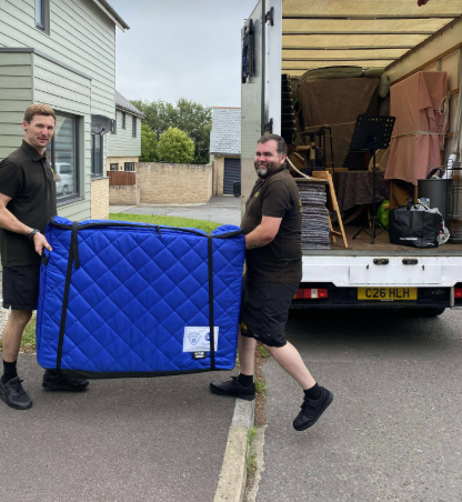Two movers carry a blue quilted sofa from a truck on a residential street