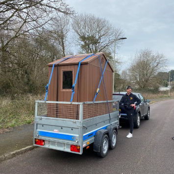 Man standing beside a car towing a small brown wooden cabin on a trailer along a road