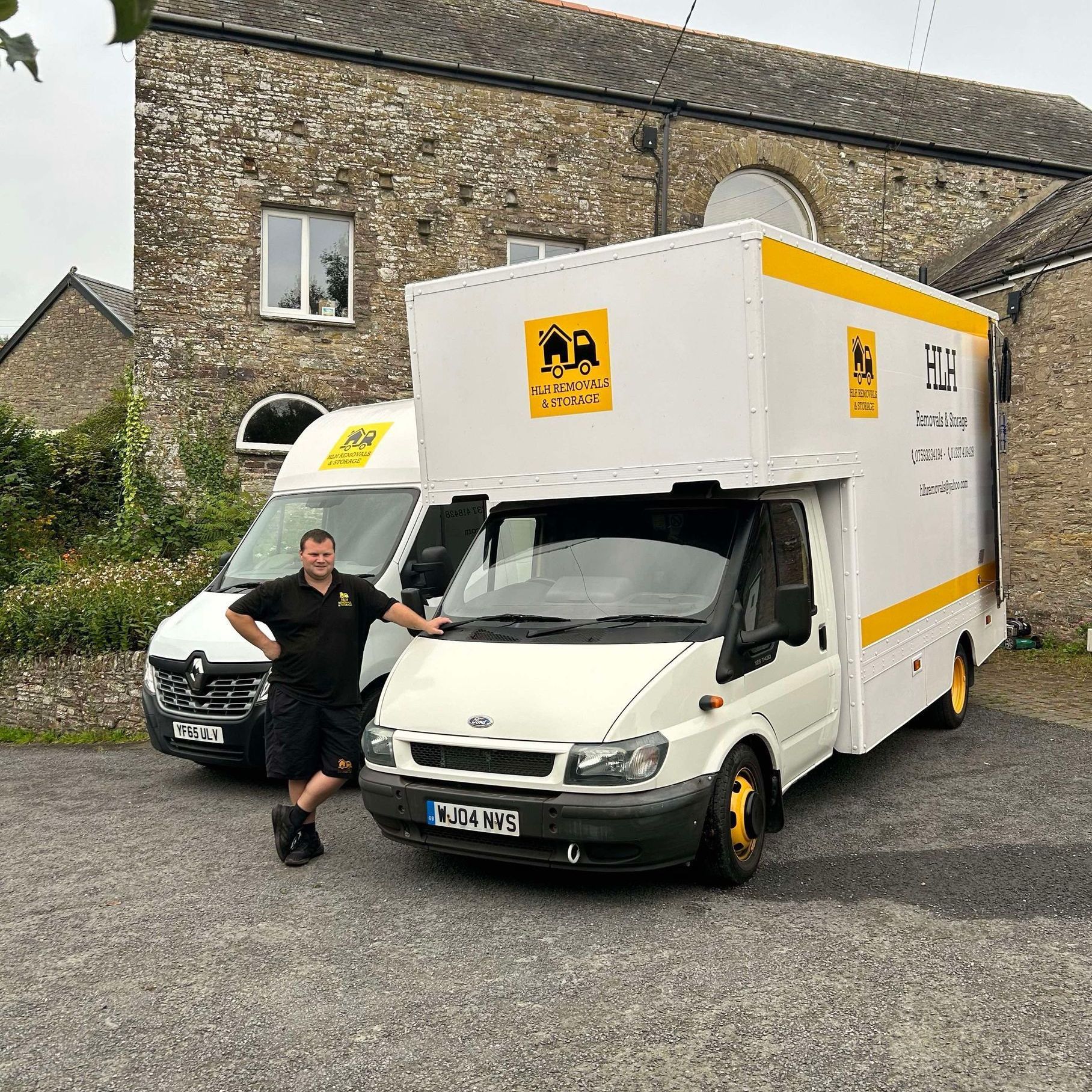 Person leaning on white moving truck and van outside a stone building, with yellow logo signs.