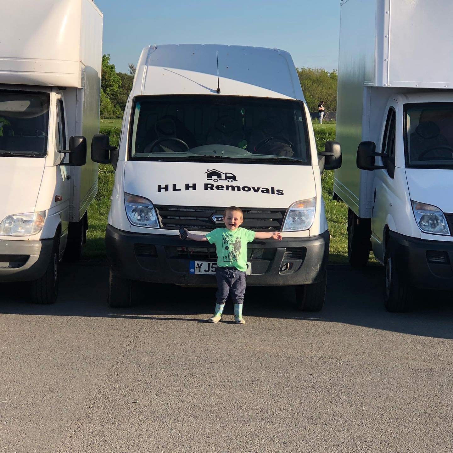 Child standing in front of a white HLH Removals van between two trucks on a sunny road