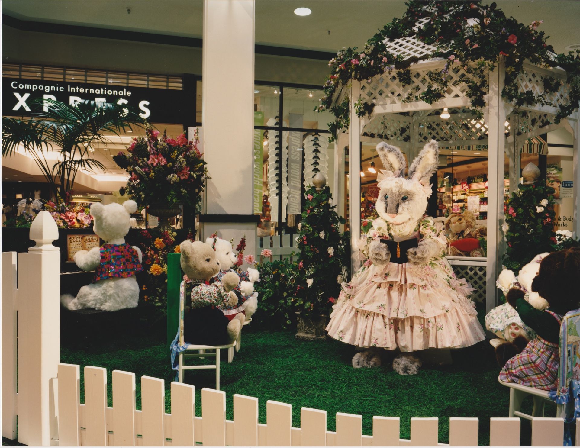 Easter display: a bunny in a pink dress stands by a gazebo in a mall, surrounded by teddy bears and flowers.