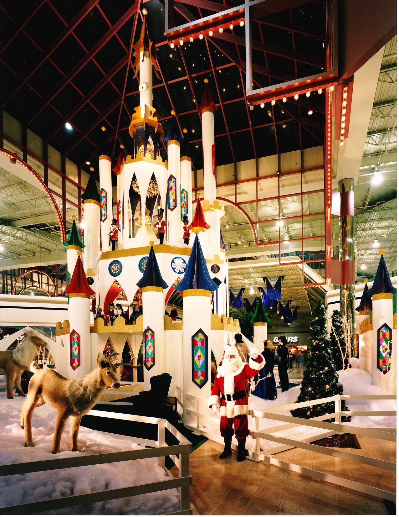 Santa stands in a festive winter scene with a large castle, snow, and colorful decorations inside a building.