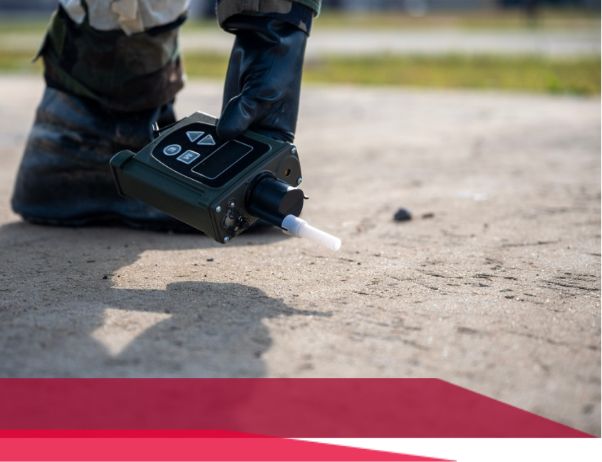 Person in military uniform uses a portable chemical detection device outdoors.