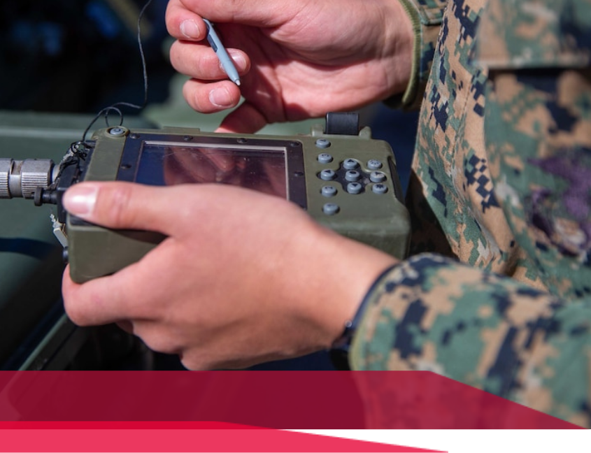 Soldier holding a rugged handheld device, possibly for communication or targeting, outdoors.