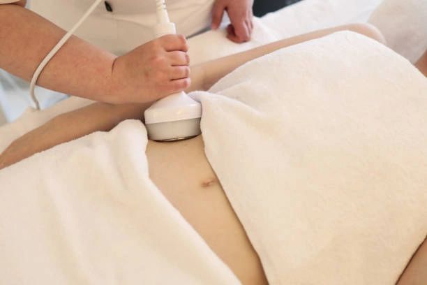 Person receiving a body treatment, device held over abdomen, surrounded by white towels.