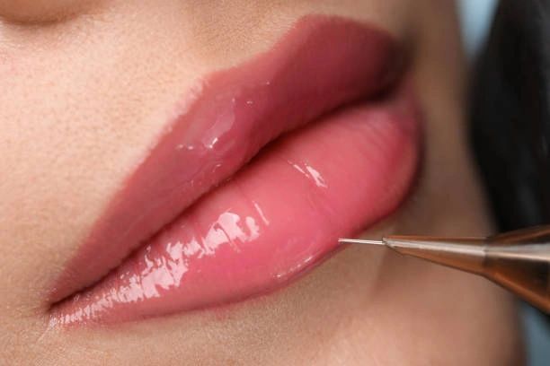 Lip tattoo being applied to full, pink lips. Close-up shows the needle and skin.