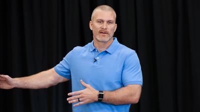 A man in a blue shirt is standing in front of a black curtain.