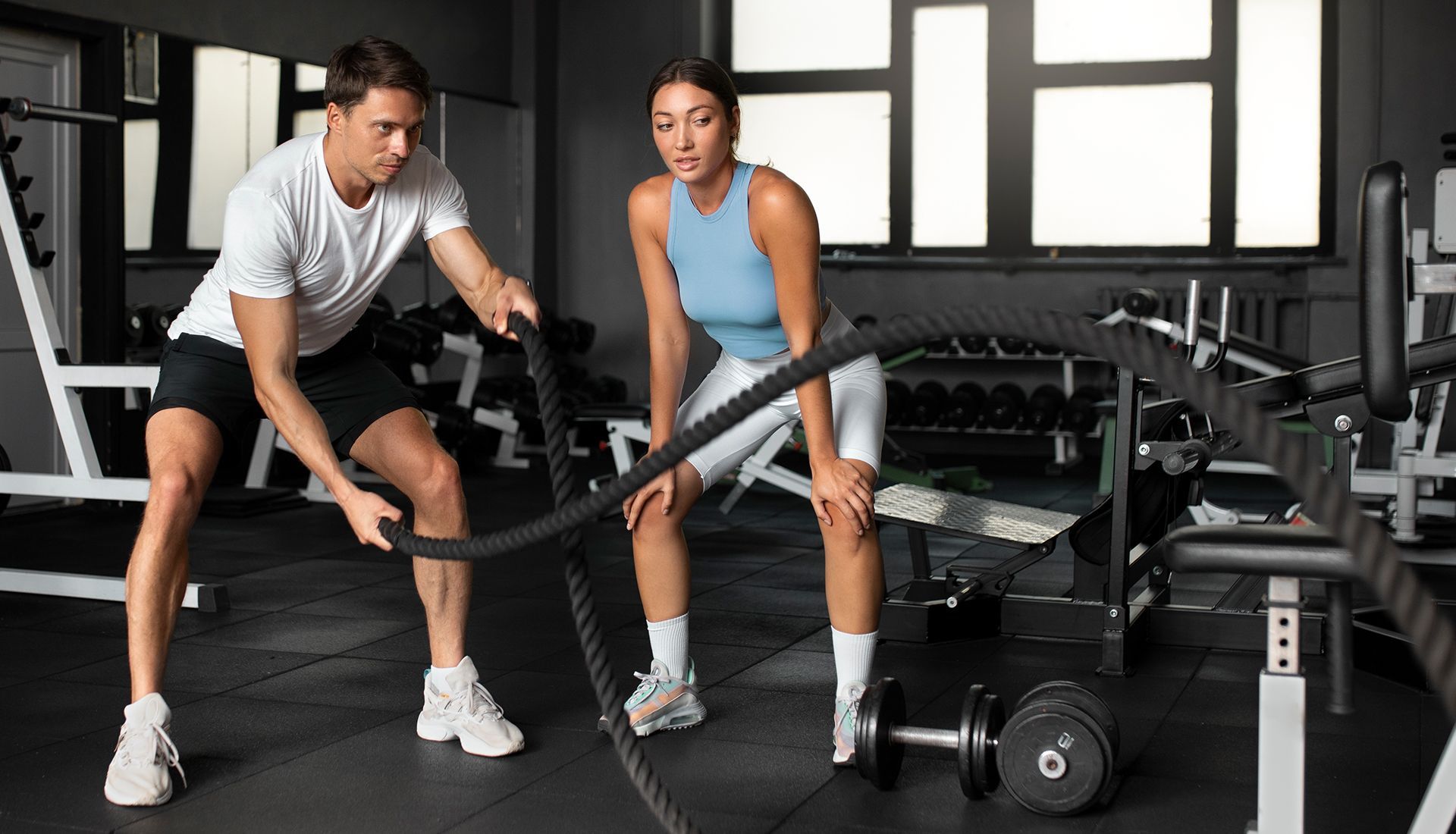 A person in a white shirt trains with battle ropes while another person watches in a modern gym.