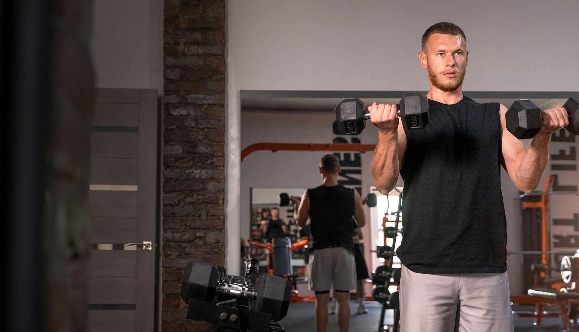 A focused athlete performs a dumbbell bicep curl in a gym, with another person visible in a mirror in the background.