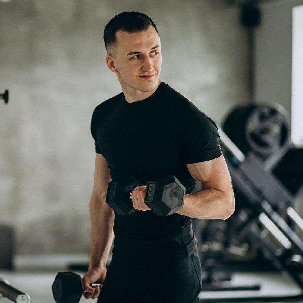A person in black athletic gear exercises with dumbbells in a gym, looking away with a smile.
