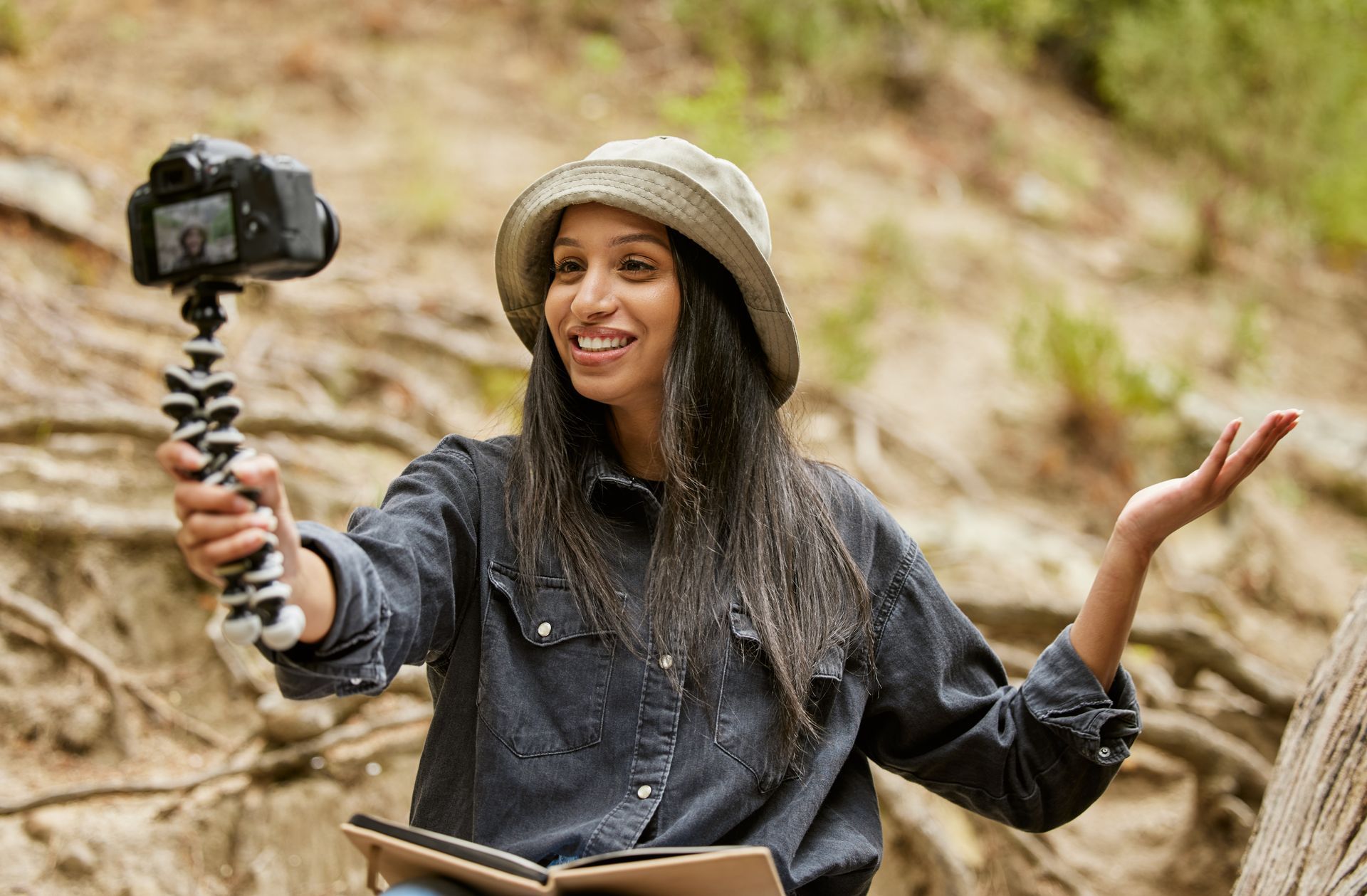 Woman in a hat holding a camera on a tripod, gesturing, talking, and smiling outdoors.