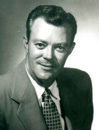 Black-and-white portrait of a man in a suit and patterned tie, smiling slightly at the camera.