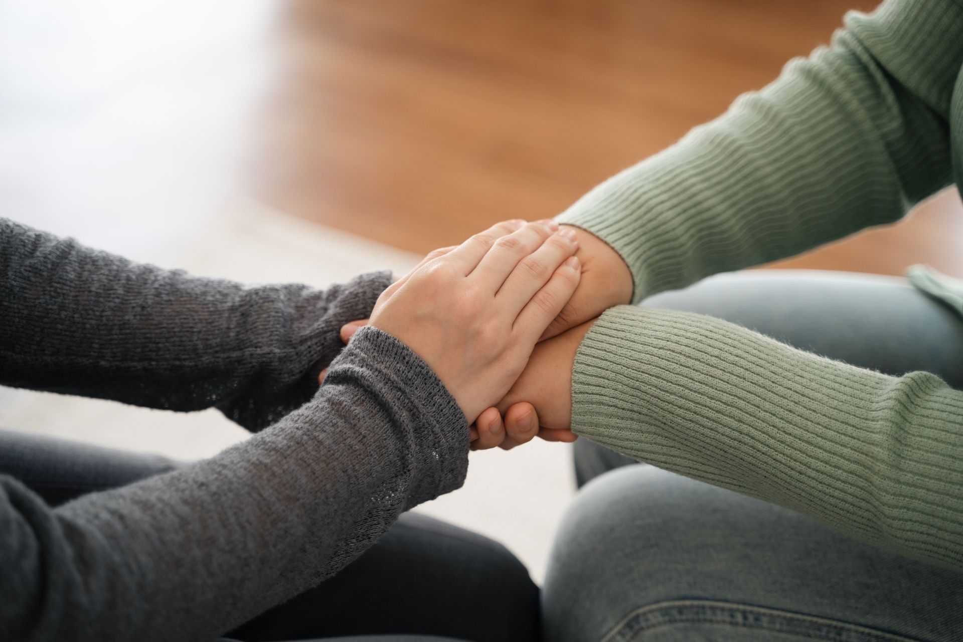 Two hands clasped across a wooden table, one wearing a watch and yellow sweater sleeve.