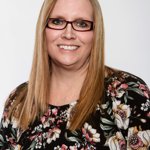 Smiling woman with glasses and long blonde hair wearing a black floral top on a white background