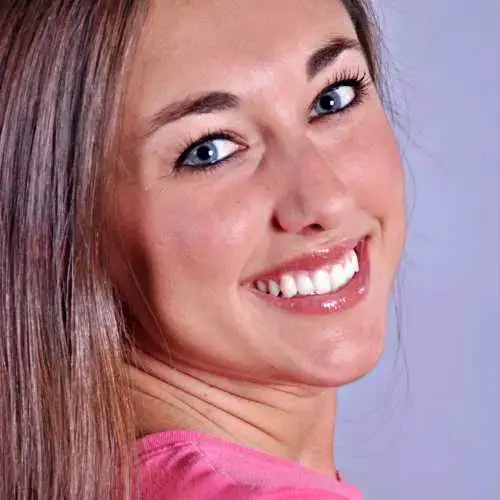 Smiling woman with blue eyes and long brown hair in a pink top, turning over her shoulder against a purple background