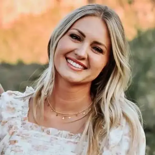 Smiling blonde woman in a white floral top outdoors with a blurred orange and green background