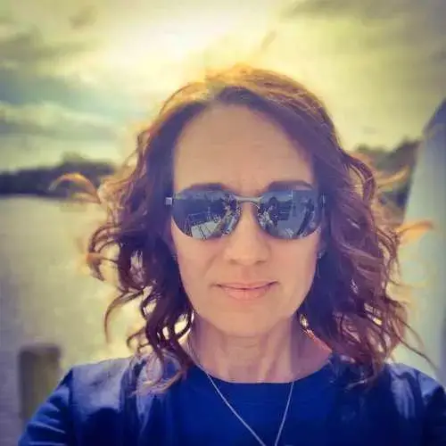 Person wearing sunglasses and a navy shirt, standing outdoors by water in bright sunlight.